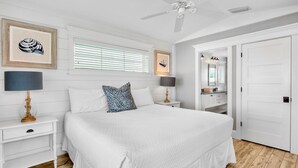 A bedroom with a large bed, two bedside tables with lamps, a ceiling fan, and a door leading to a bathroom.