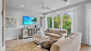 A living room with a sofa, a coffee table, a TV, and a view of greenery through large windows.