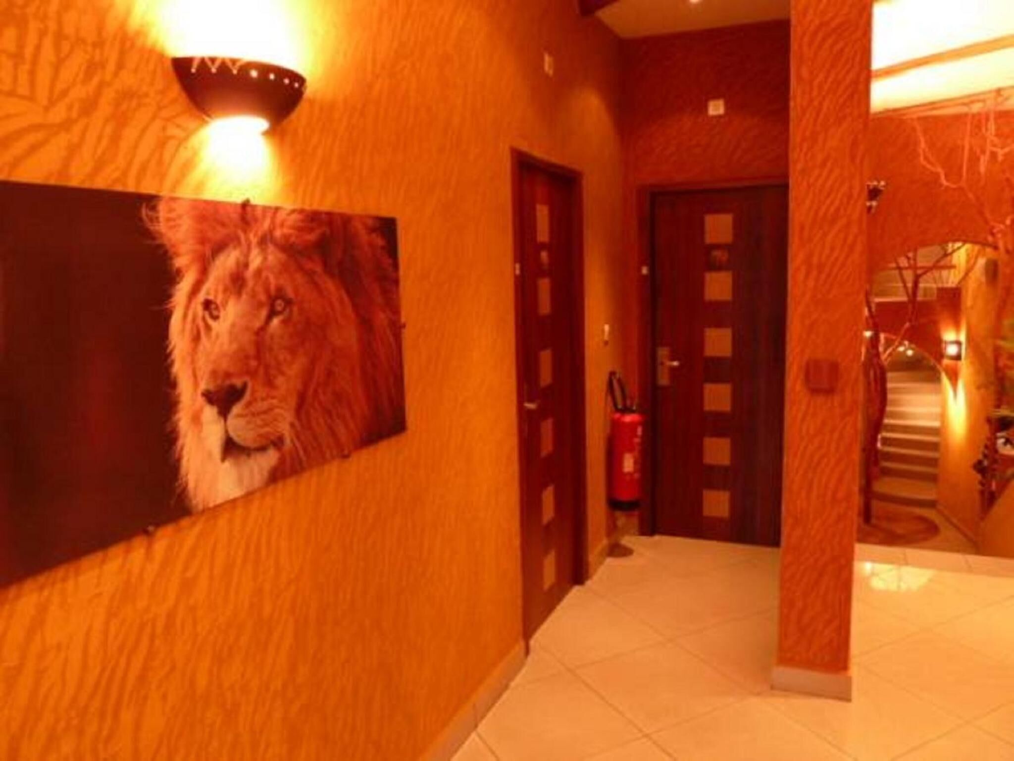 A hallway with a wooden floor, a framed picture of a lion, a red fire extinguisher, and a staircase in the background.