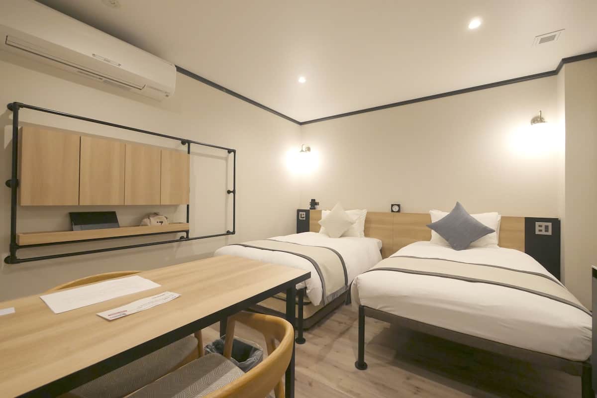 Habitación de hotel con dos camas, un escritorio, una silla, una mesita, un estante y un equipo de aire acondicionado.