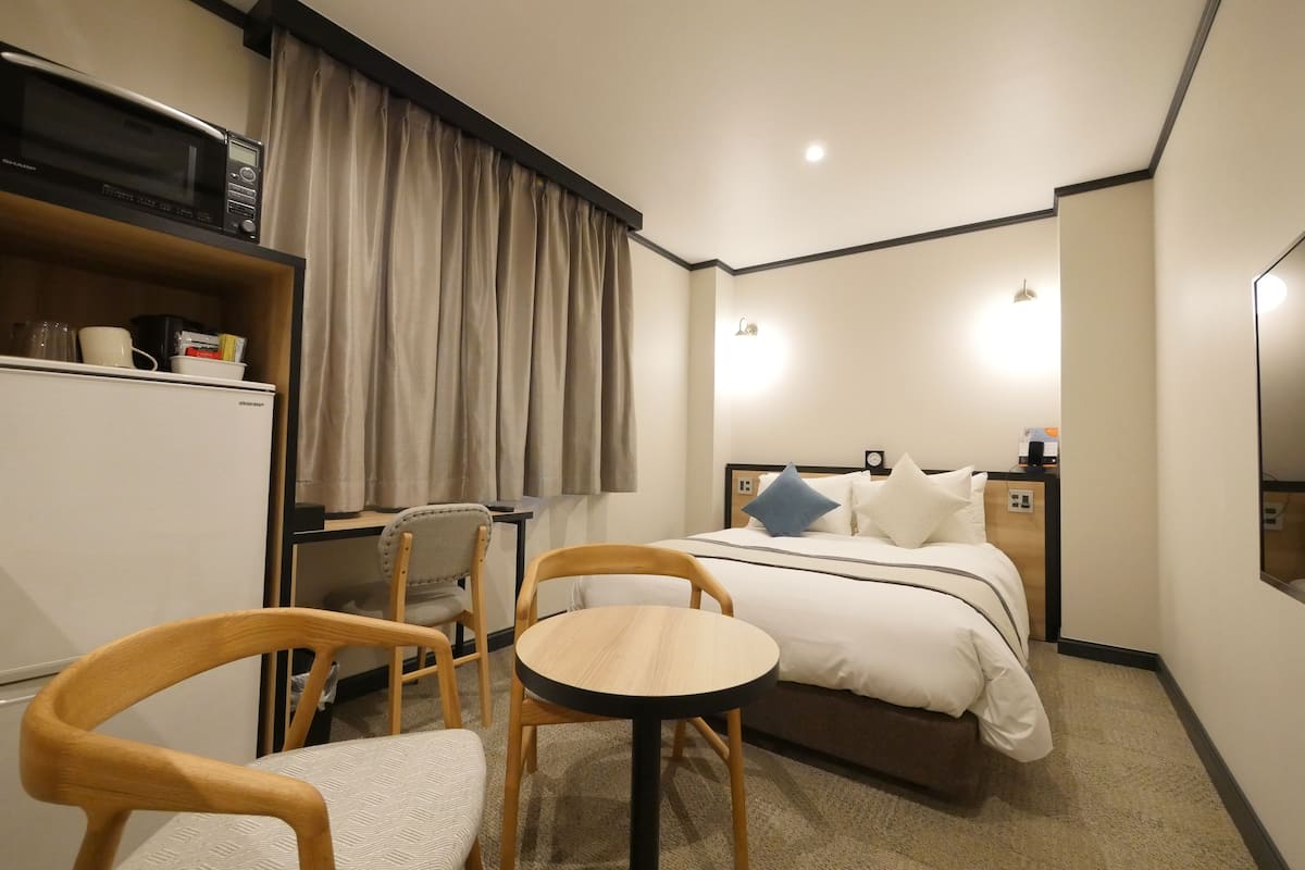Habitación de hotel con cama, mesita de noche, una pequeña mesa redonda, una silla, escritorio, microondas y televisión.