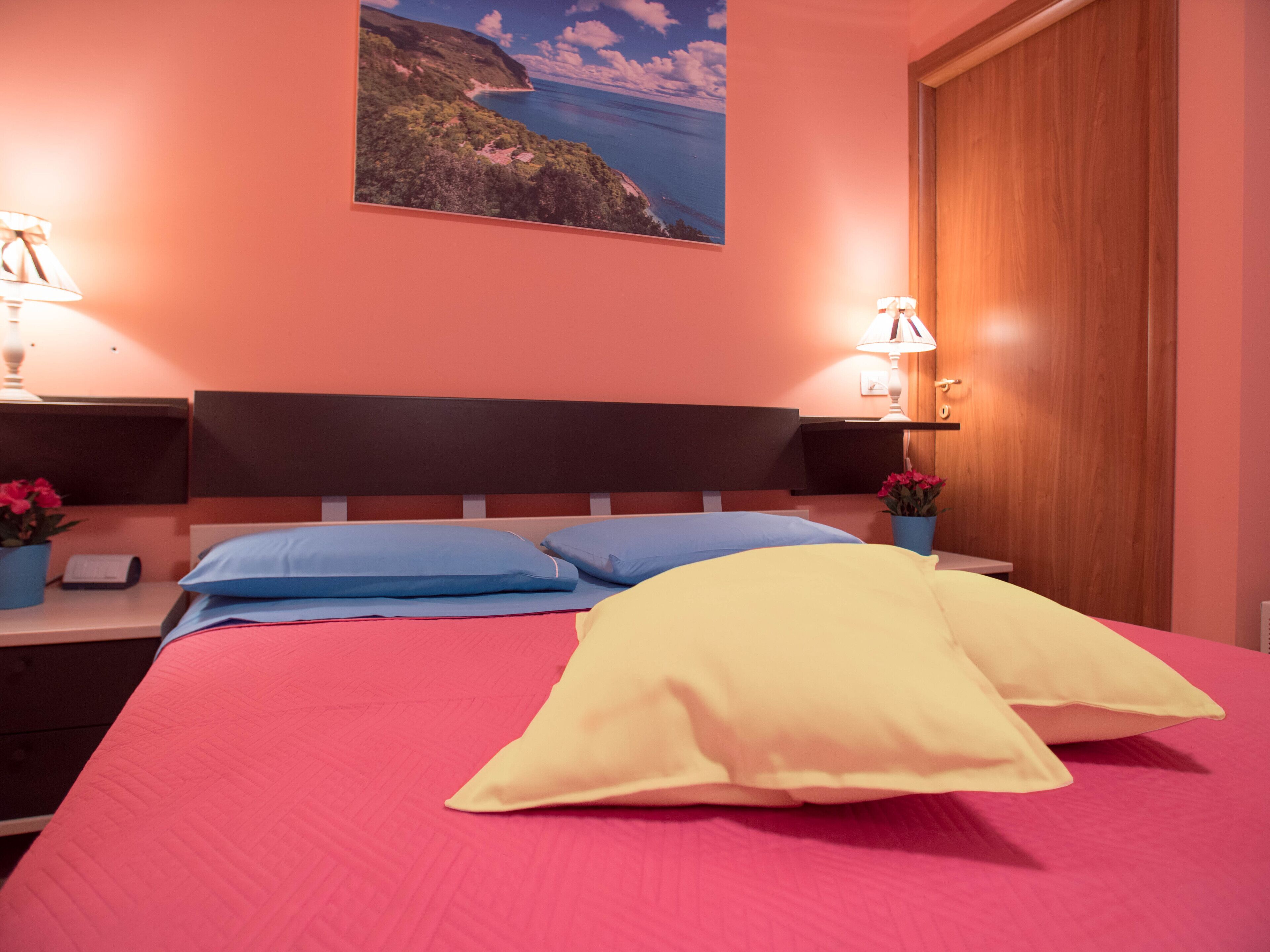 Un dormitorio con una cama, dos mesitas de noche, una mesita auxiliar, una puerta de madera y un cuadro colgado en la pared.