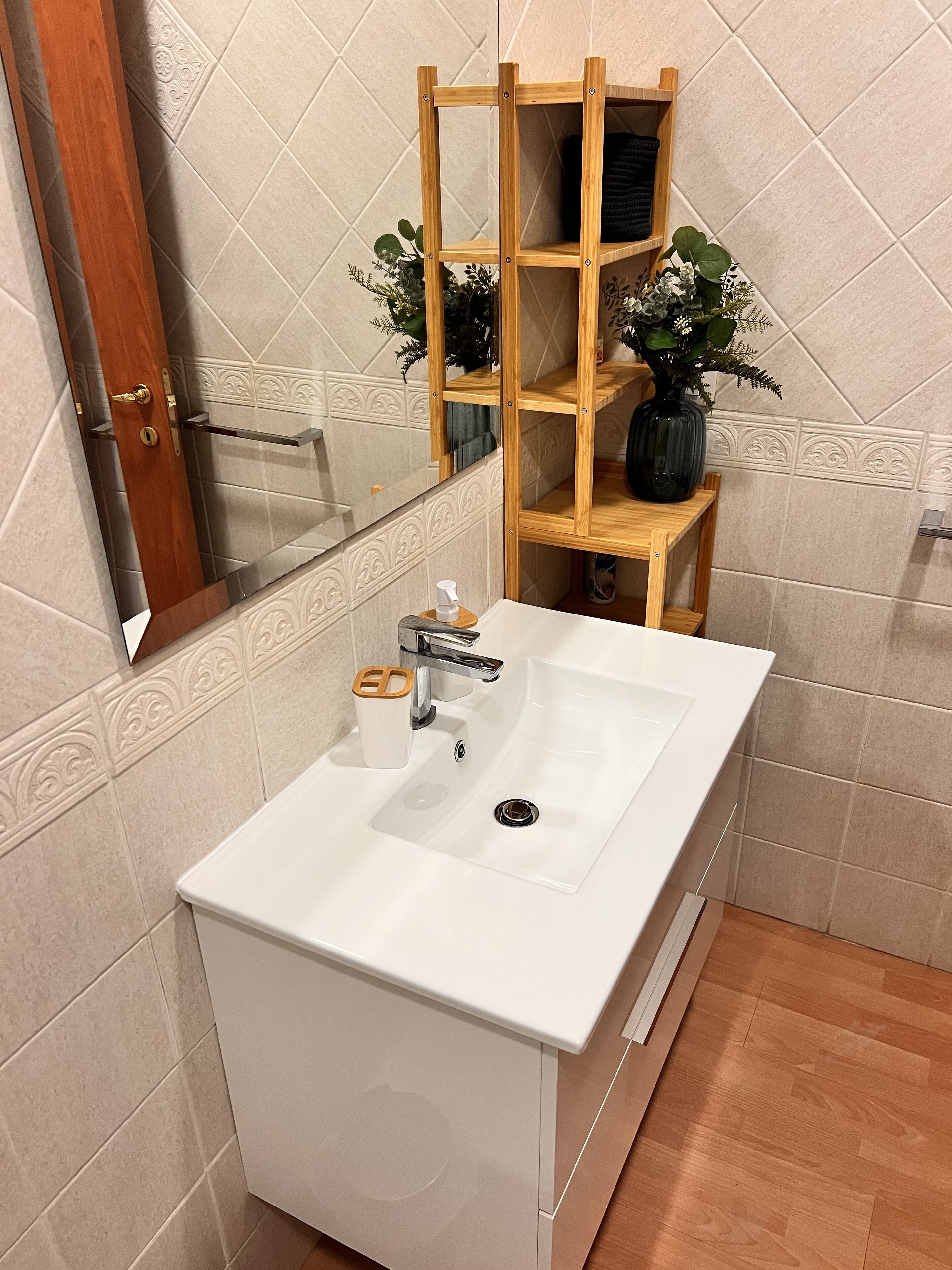 Ett badrum med ett vitt handfat, hyllor i trä, en spegel och en dörr.