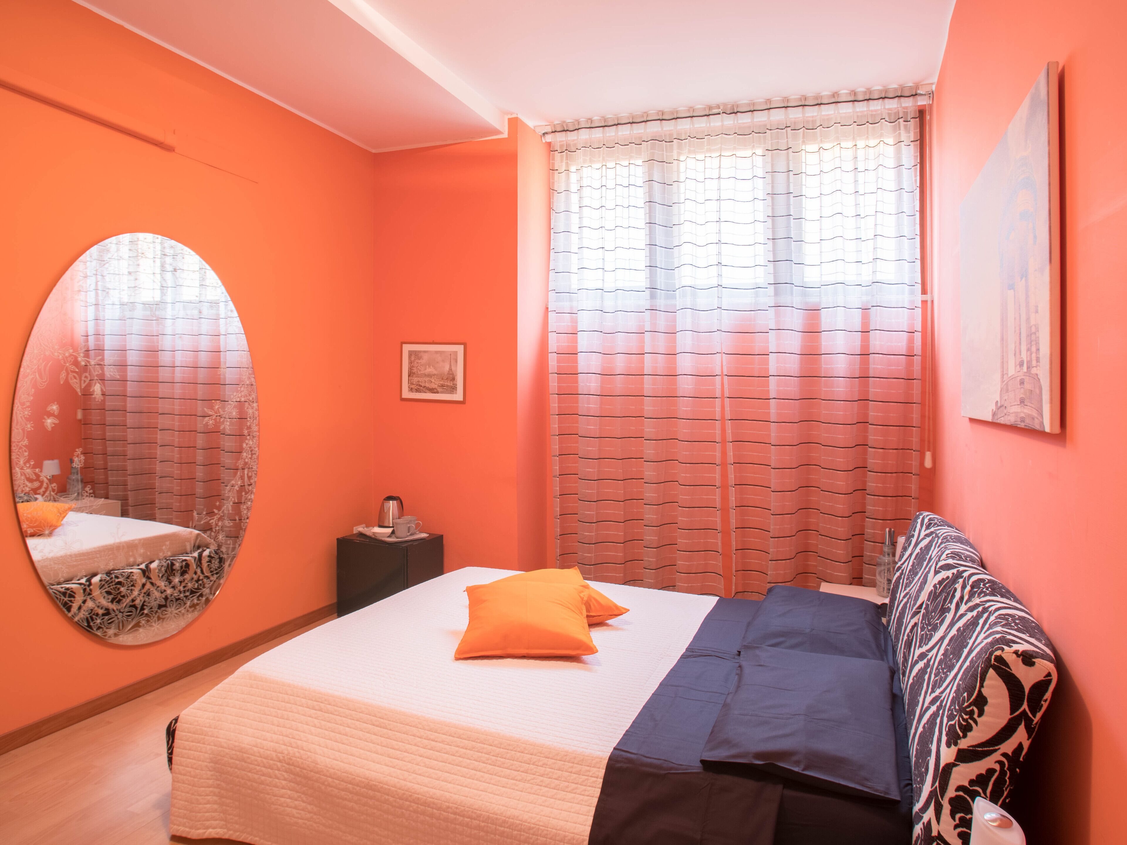 Ein Schlafzimmer mit einem Bett, einem runden Spiegel, einem Nachttisch mit Lampe und einem Fenster mit Vorhängen.