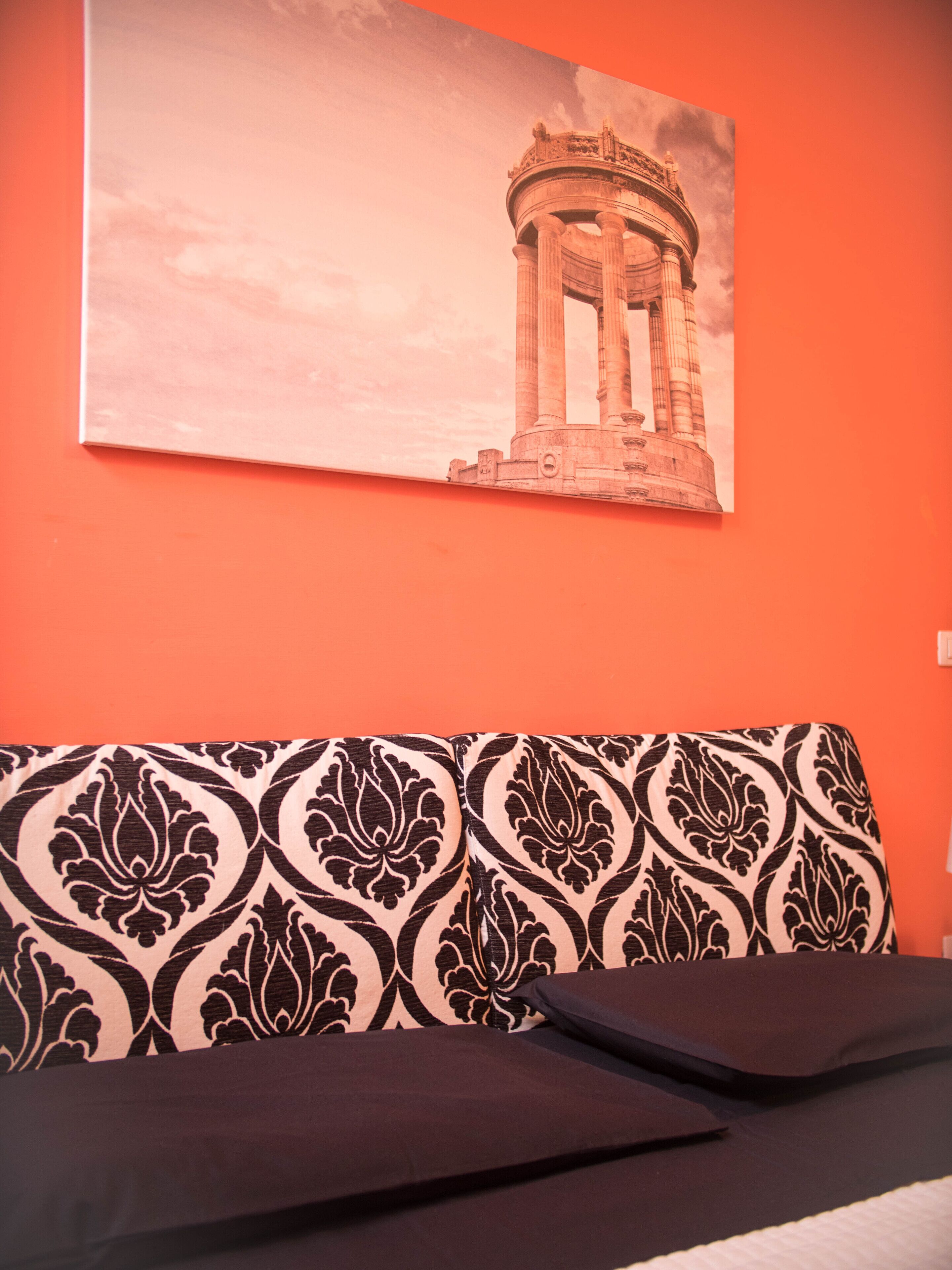 A room with a patterned sofa, a bed with a black headboard, and a wall-mounted painting of a classical building.