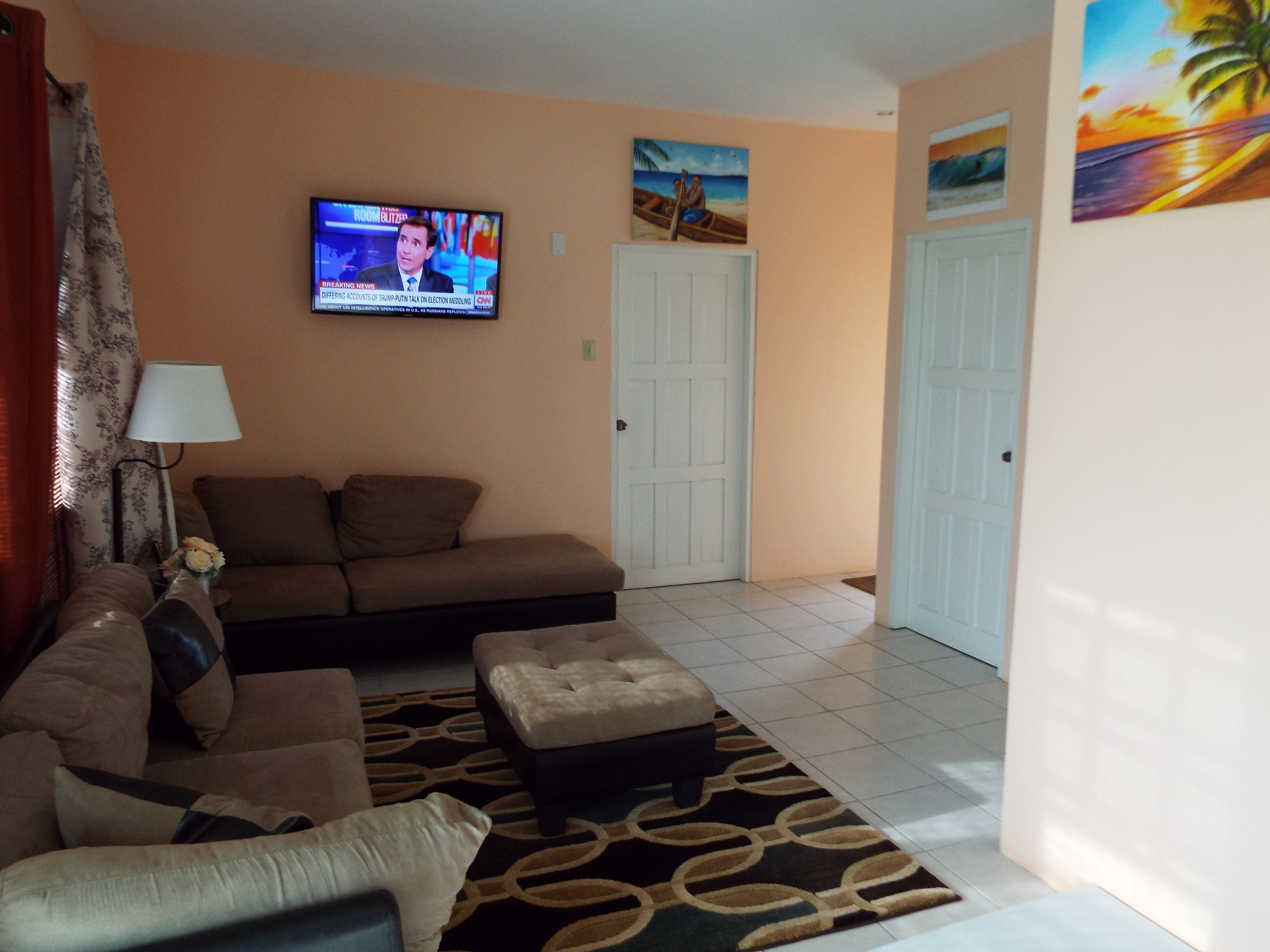 A living room with a sofa, ottoman, and a TV displaying a news channel.