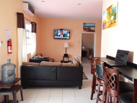 Una sala de estar con un sofá negro, un televisor, una laptop sobre un escritorio y un cuadro en la pared.