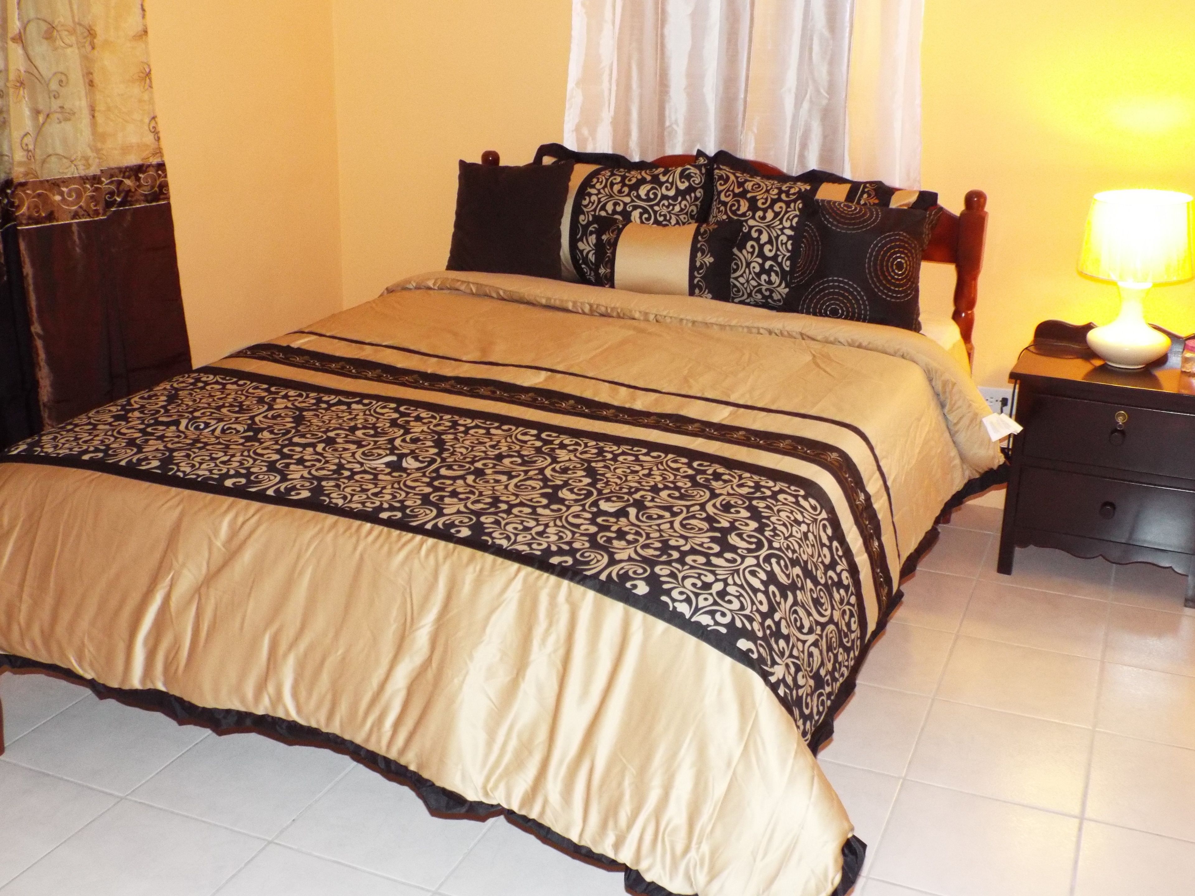 A neatly made bed with a patterned bedspread and pillows, a bedside table with a lamp, and a curtain on the window.