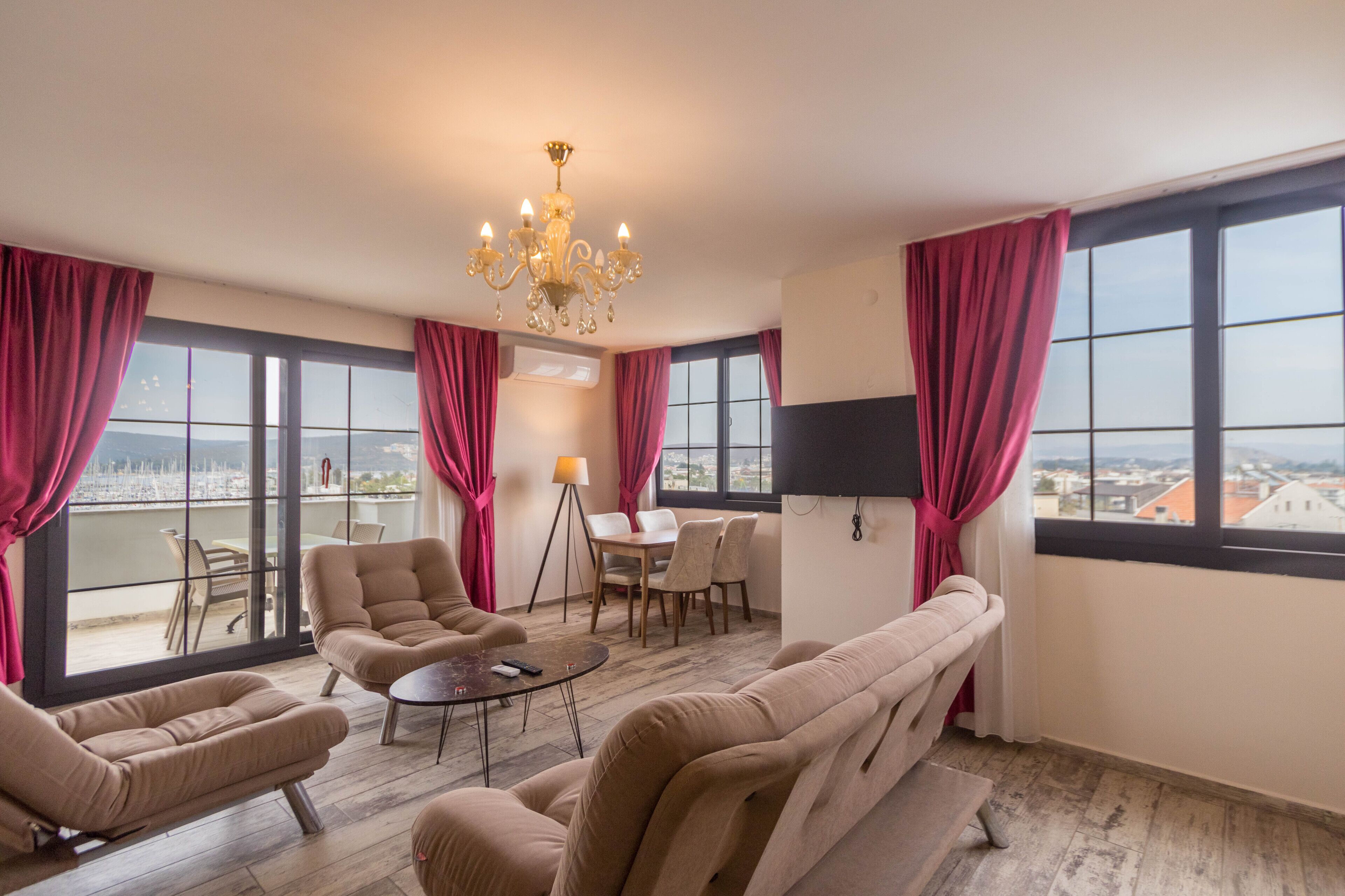 Een ruime woonkamer met een bank, fauteuils, een salontafel en een kroonluchter. Er is een eethoek met een tafel en stoelen, en grote ramen met gordijnen.