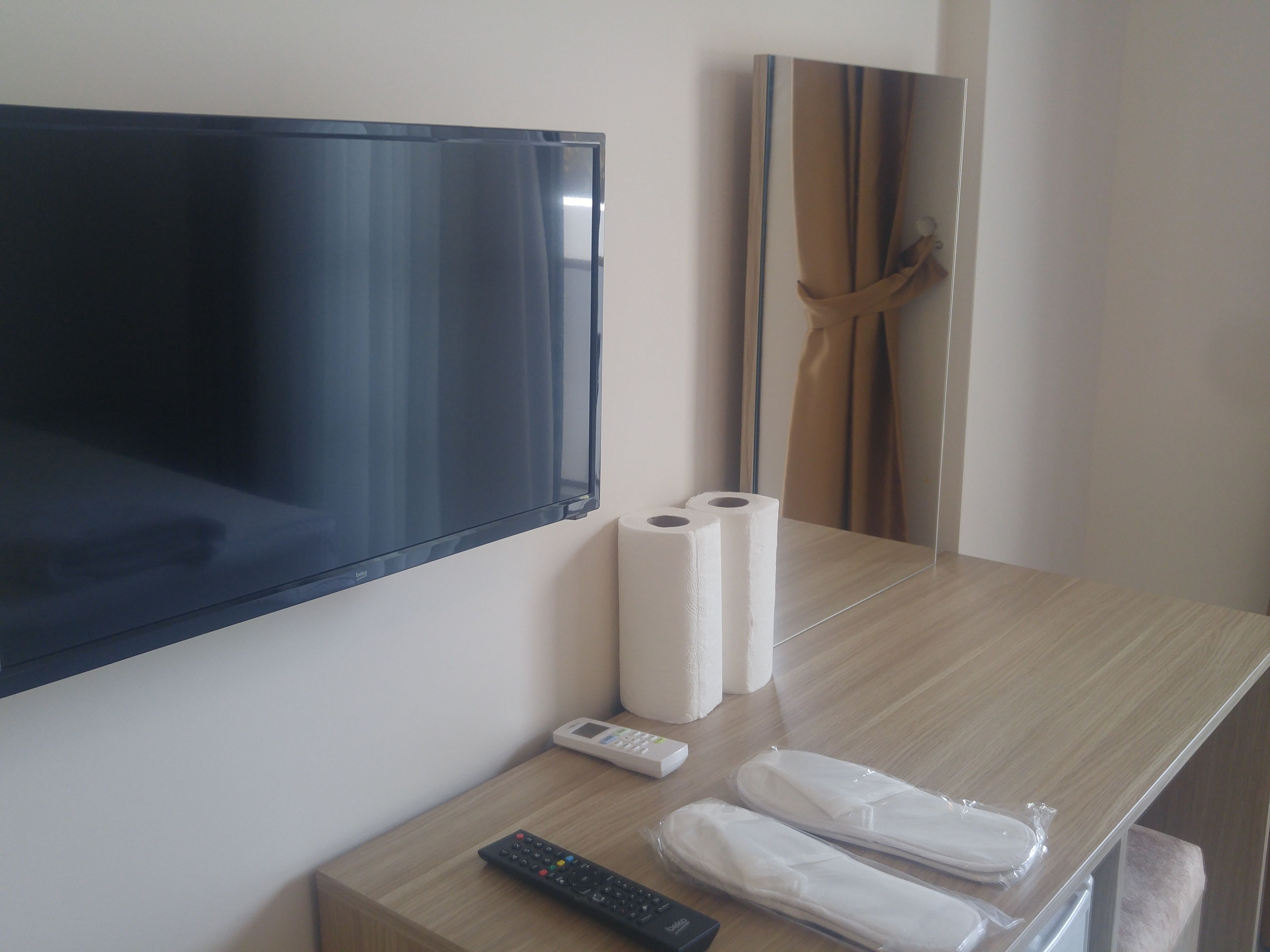 A hotel room with a mounted TV, two rolls of toilet paper, a remote control, and a pair of slippers on a wooden nightstand.