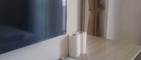 Habitación de hotel con un televisor de pared, dos rollos de papel higiénico, un control remoto y un par de pantuflas sobre una mesita de noche de madera.