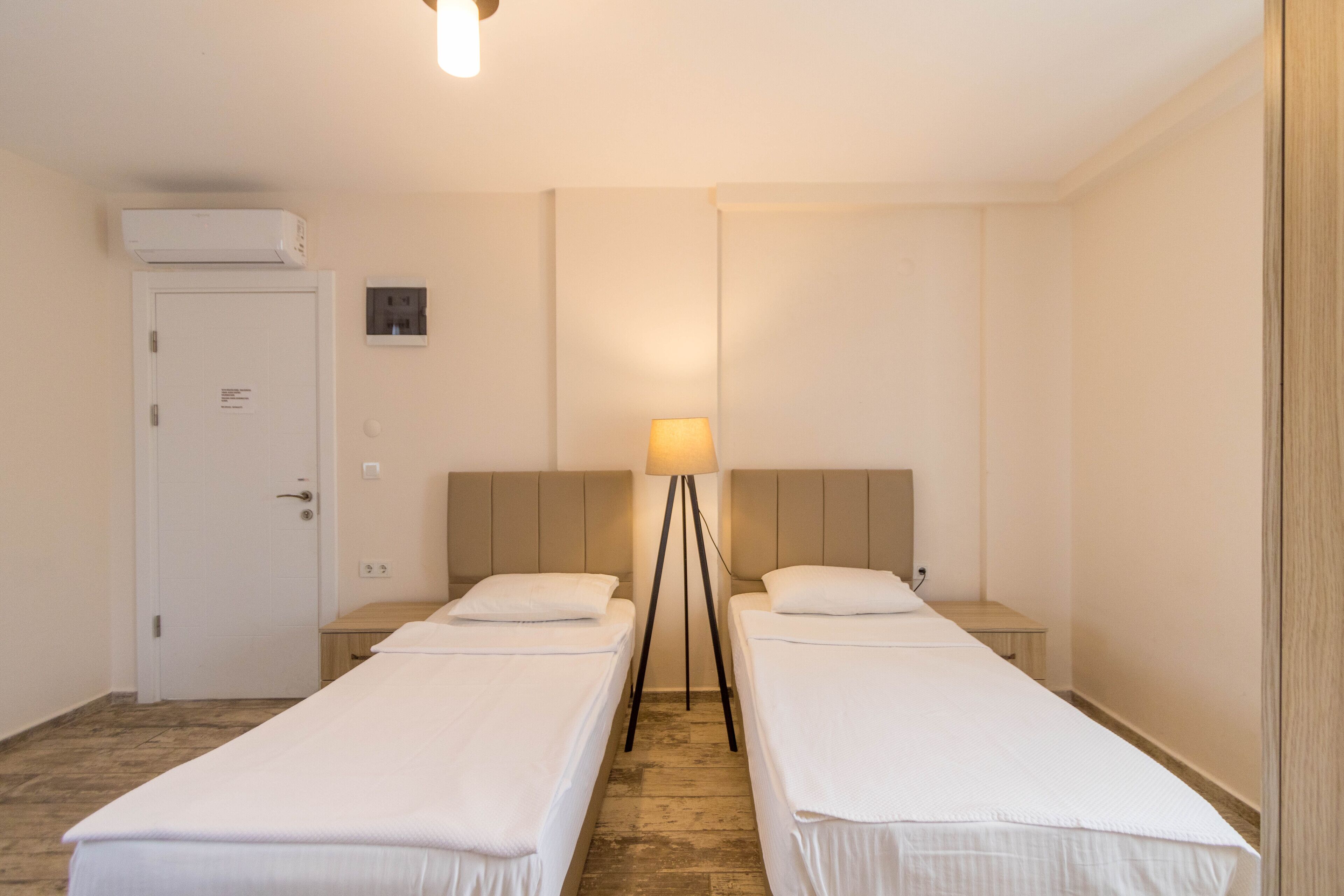 Habitación de hotel con dos camas individuales, una mesita de noche con lámpara, un armario y un equipo de aire acondicionado.
