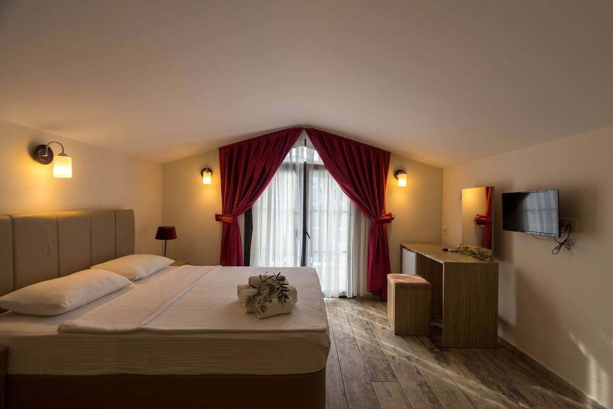 A hotel room with a large bed, a desk, a television, and a window with red curtains.