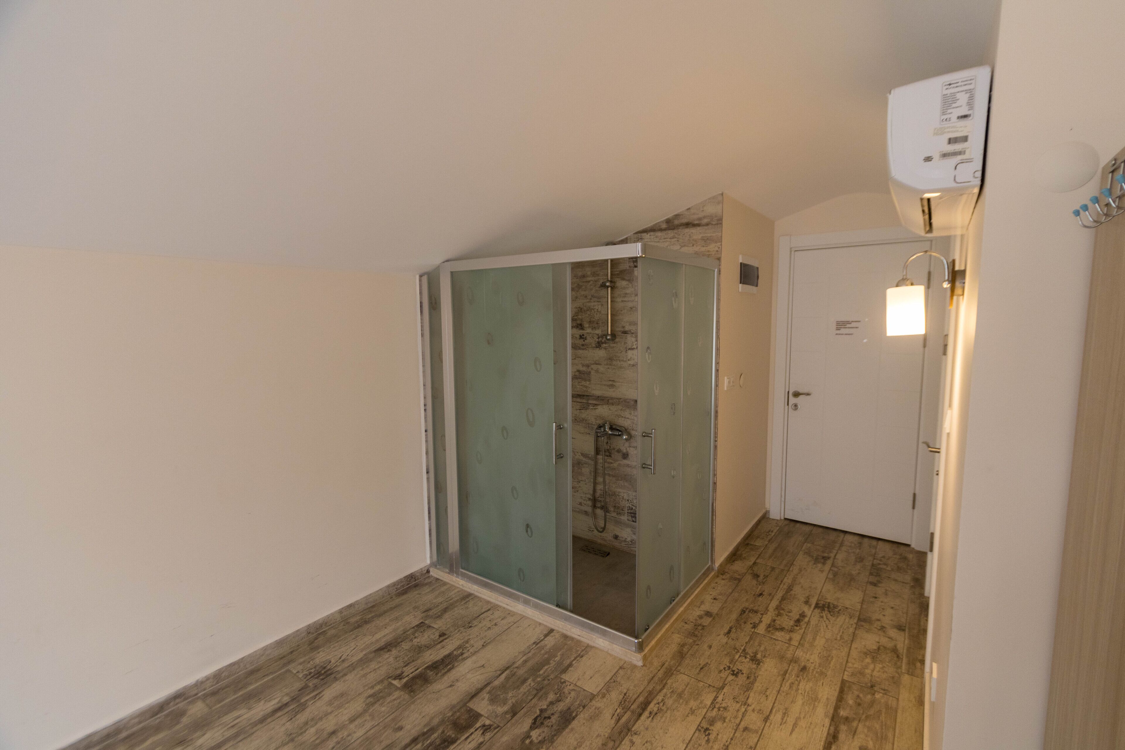 Een kamer met een douche in een glazen cabine, houten vloer, een witte deur en een wandairconditioner.