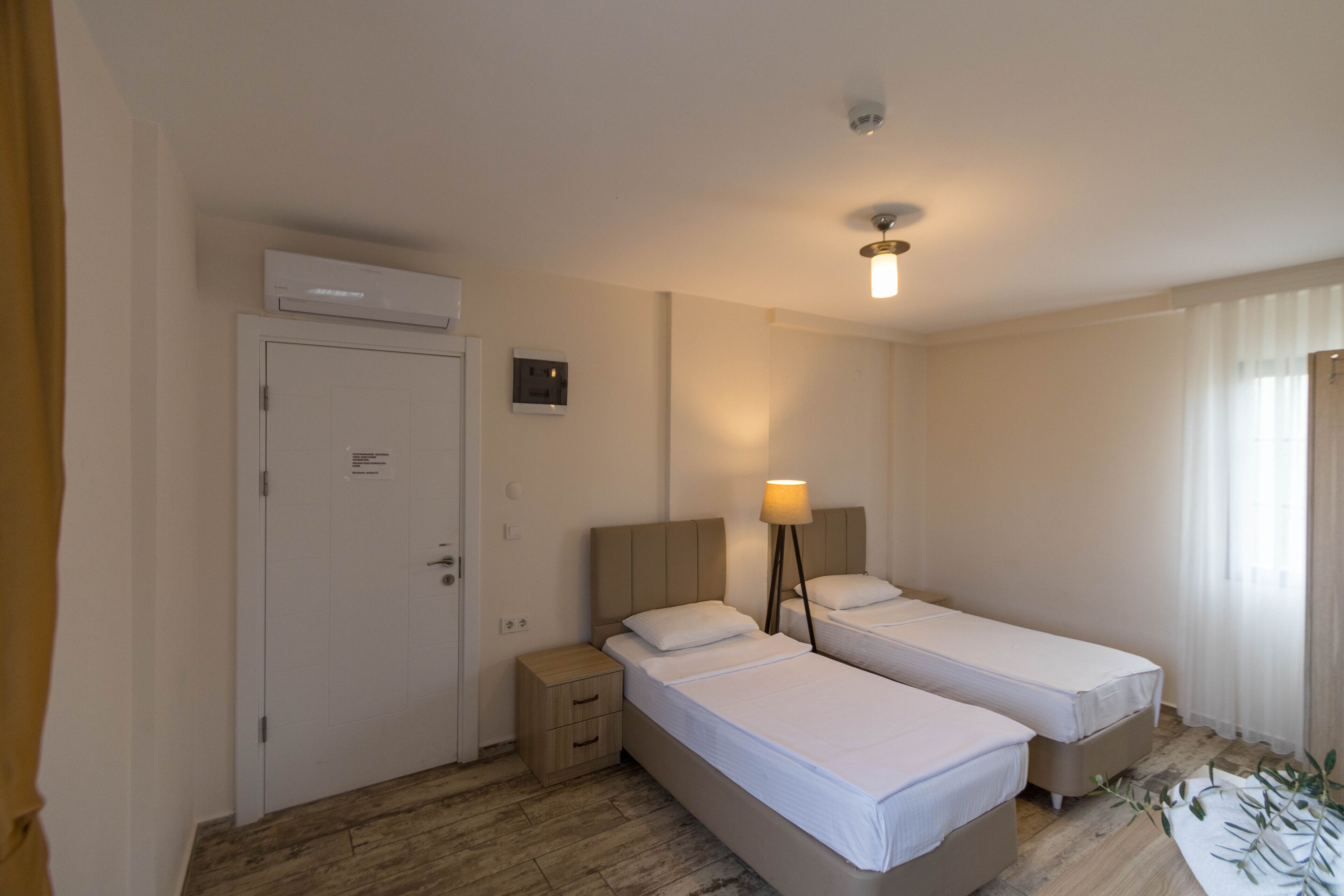 A hotel room with two single beds, a bedside table, a nightstand, an air conditioning unit, and a ceiling light.