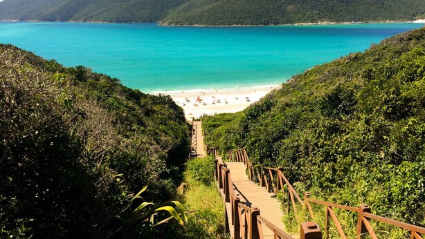 A wooden staircase leading to a beach with turquoise water and green hills in the background. - Apartment in the city center (Praia dos Anjos)