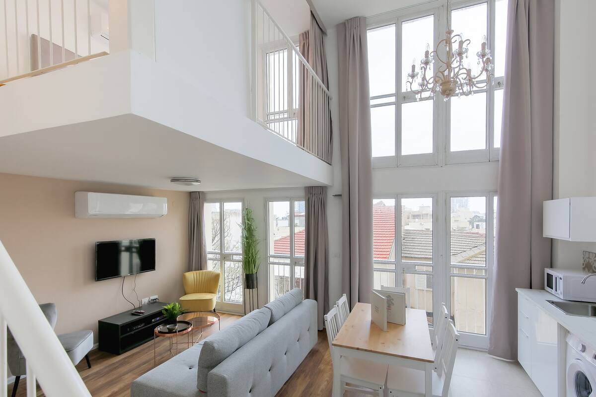 A modern apartment with a loft bedroom, a dining area, a kitchen, and a living room with a grey sofa and a TV.