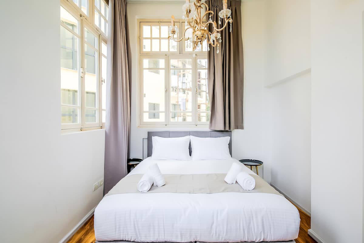 A neatly made bed with white linens and two rolled towels, a chandelier, and a view of buildings through large windows.