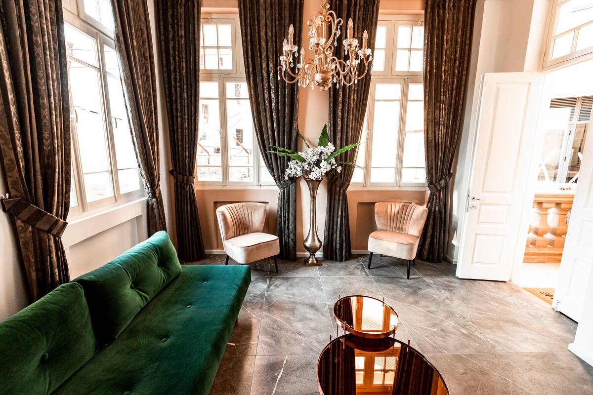 A spacious room with a green sofa, two armchairs, a tall vase with flowers, and a chandelier.