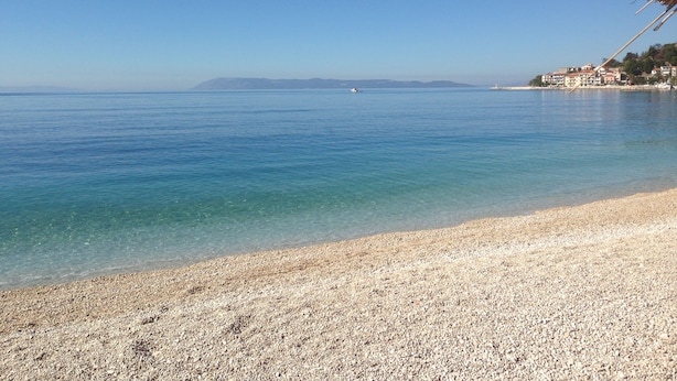 A beach with clear water, pebbly shore, and a distant town.
