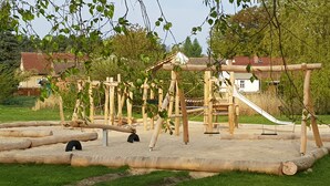 Ein Spielplatz mit Holzkonstruktionen, darunter Schaukeln und eine Rutsche, inmitten einer grünen Fläche mit Bäumen und Häusern im Hintergrund.
