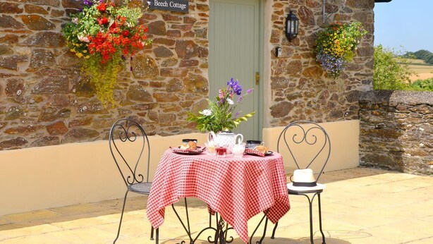 A stone cottage with a checkered tablecloth, a sign reading "Beech Cottage," and flower baskets on the wall. - Rural Complex 7 Lux Cottages, Beech Cottage Sleeps 2, 1 well behaved dog allowed (Tregony)