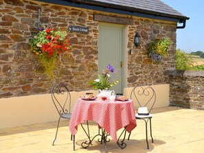 Une petite maison en pierre avec une nappe à carreaux, une enseigne indiquant « Beech Cottage », et des jardinières suspendues au mur.