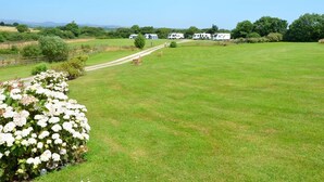 A spacious grassy field with a clear sky, surrounded by trees and a few white RVs parked in the distance. - Rural Complex 7 Lux Cottages, Beech Cottage Sleeps 2, 1 well behaved dog allowed (Tregony)