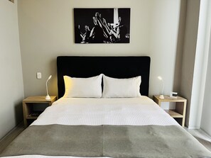 A neatly made bed with white linens and a grey throw blanket, flanked by two bedside tables with lamps, in a modern room with a black headboard and a black and white artwork above. - Hotel Arena Suites Bogota (Bogotá)