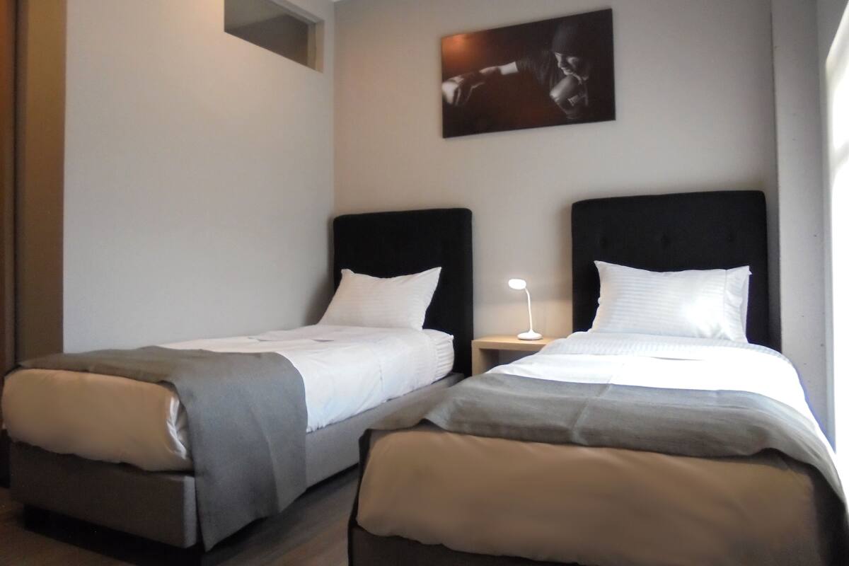 Deux lits simples avec des têtes de lit noires, une petite table de chevet avec une lampe et un tableau encadré au mur.