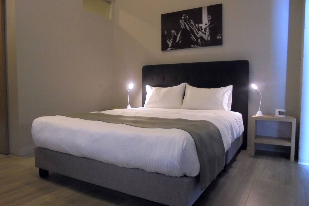 Une chambre à coucher moderne avec un grand lit, deux lampes de chevet, une tête de lit et une œuvre d’art encadrée accrochée au mur.