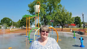 Una mujer está de pie en un parque acuático con diversas estructuras de juego y un cielo azul despejado.