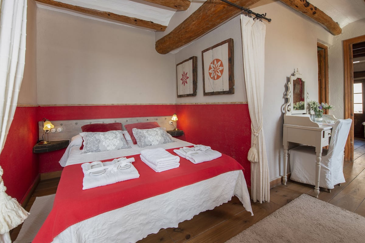 Quarto com esquema de cores vermelho e branco, vigas de madeira, cama com roupa de cama branca, mesa de cabeceira com abajur e um espelho.
