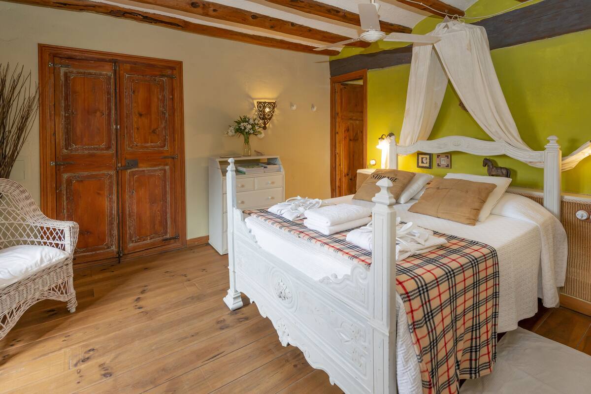 Quarto com cama de dossel, guarda-roupa de madeira, cadeira de vime e mesa de cabeceira com abajur.