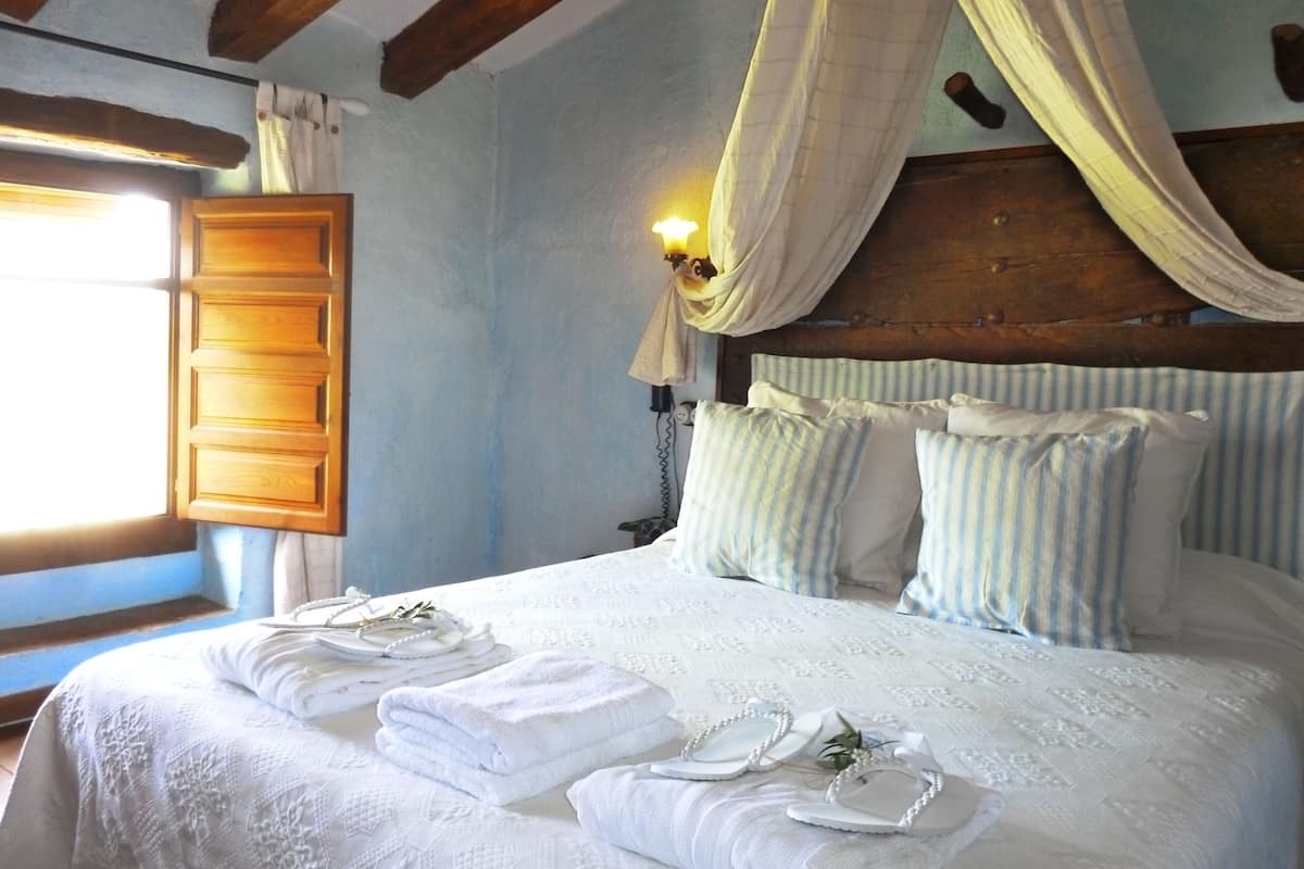 Quarto com cama, almofadas, dossel, mesa de cabeceira e janela com cortinas.