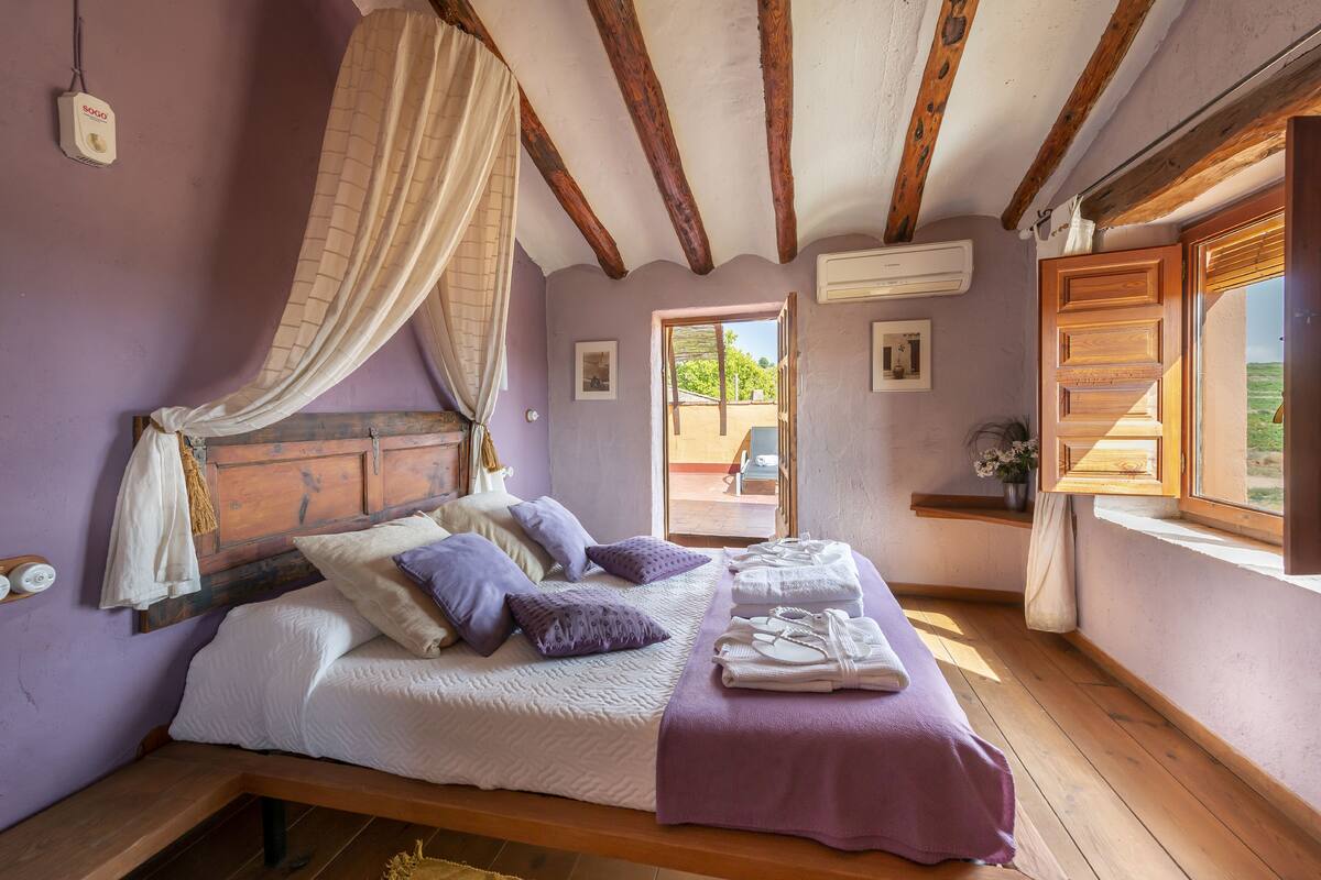 Quarto com cama de madeira, roupa de cama roxa, um dossel e uma janela com persianas de madeira.
