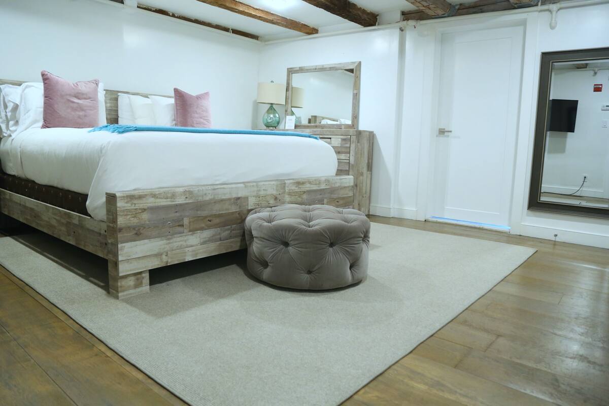 A bedroom with a wooden bed, a round ottoman, a mirror, and a white door.