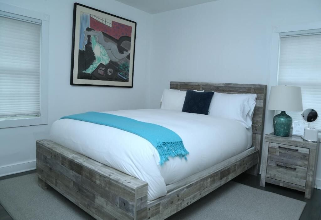 A bedroom with a wooden bed frame, white bedding, a blue throw, a nightstand with a lamp, and a framed picture on the wall.