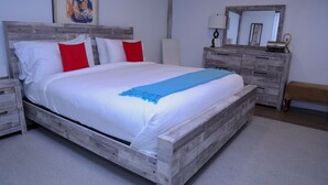 A wooden bed with white and red pillows, a blue throw, and a nightstand with drawers. - Harpoon House (Southampton)