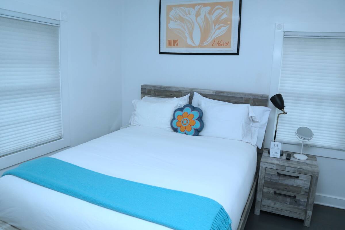A neatly made bed with a blue throw blanket, a wooden nightstand with a mirror and a small sign, and a framed picture of tulips on the wall.