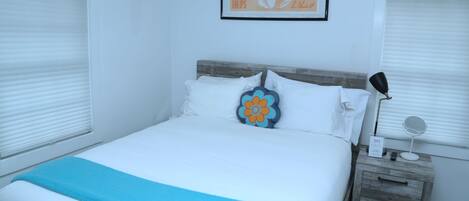 A neatly made bed with a blue throw blanket, a wooden nightstand with a mirror and a small sign, and a framed picture of tulips on the wall.