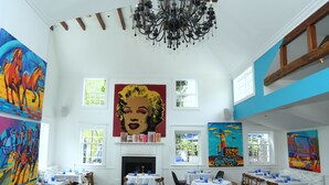 A dining area with tables set for guests, a fireplace, and large colorful paintings on the walls. - Harpoon House (Southampton)