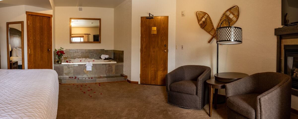 A hotel room with a bed, a bathroom with a bathtub, two armchairs, a small table, and a floor lamp.