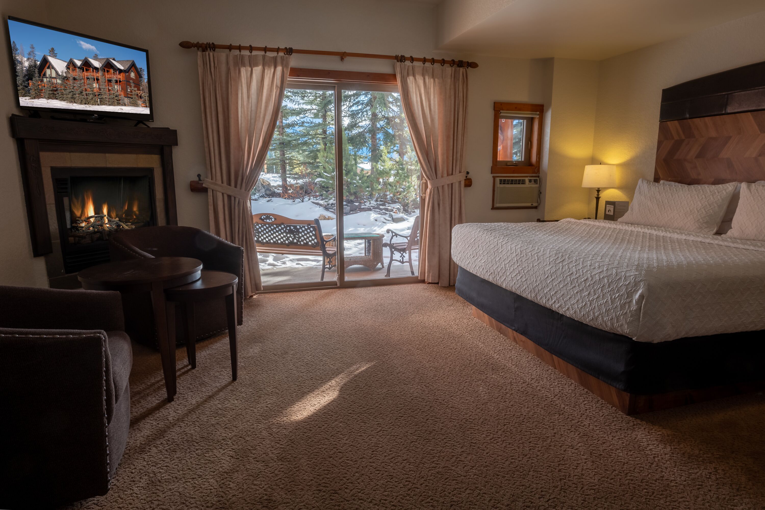 Une chambre d’hôtel avec une cheminée, un grand lit, une télévision et une vue sur un paysage enneigé, visible à travers la porte vitrée.