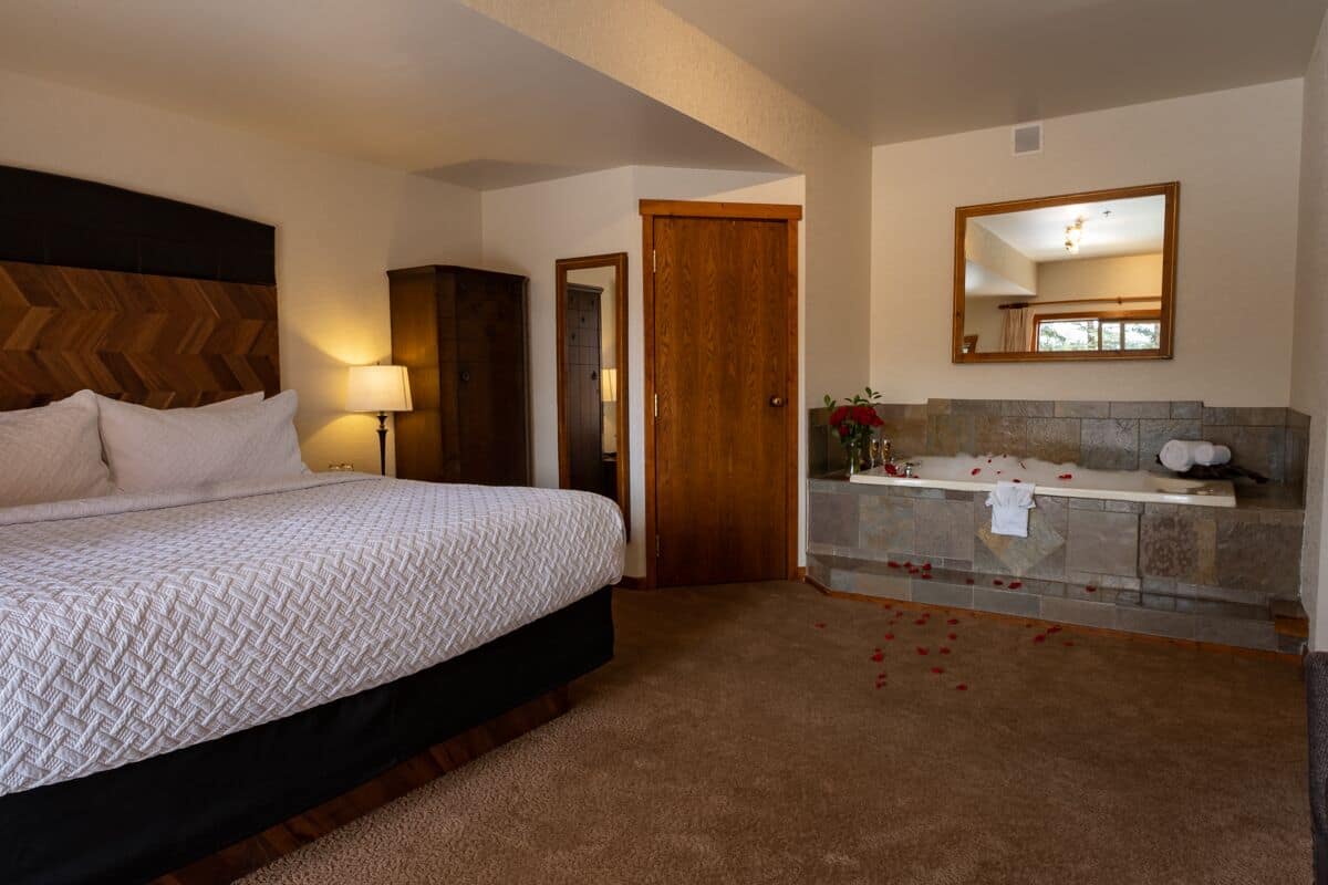 A bedroom with a large bed, a wooden headboard, a nightstand, a lamp, a bathroom with a stone sink, a mirror, and a shower area.