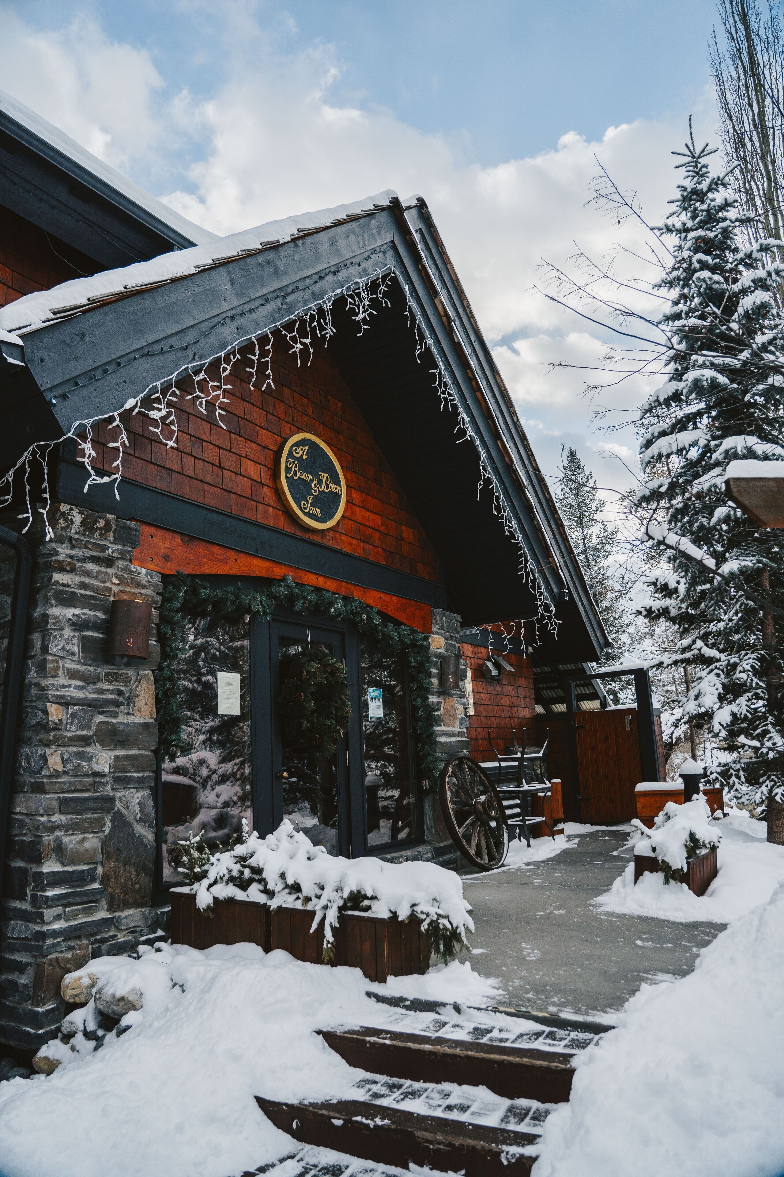 A rustic building with a stone facade, a wooden door, and a sign that reads "A Beer House Inn." Snow covers the ground and trees, and a wooden bench is visible outside.