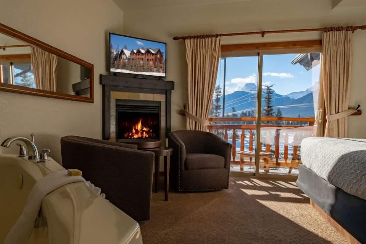 A hotel room with a fireplace, a television, a sofa, a bed, and a balcony with a mountain view.