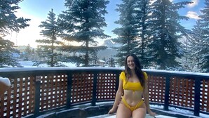 Une personne vêtue d’un maillot de bain jaune est assise au bord d’un jacuzzi, entourée d’arbres couverts de neige et d’une clôture en bois.