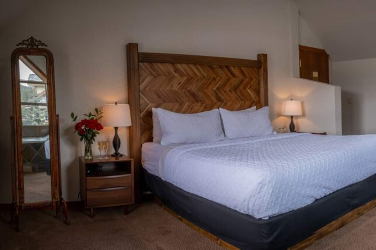 A bedroom with a large bed, bedside lamps, a mirror, and a bathroom with a bathtub and sink.