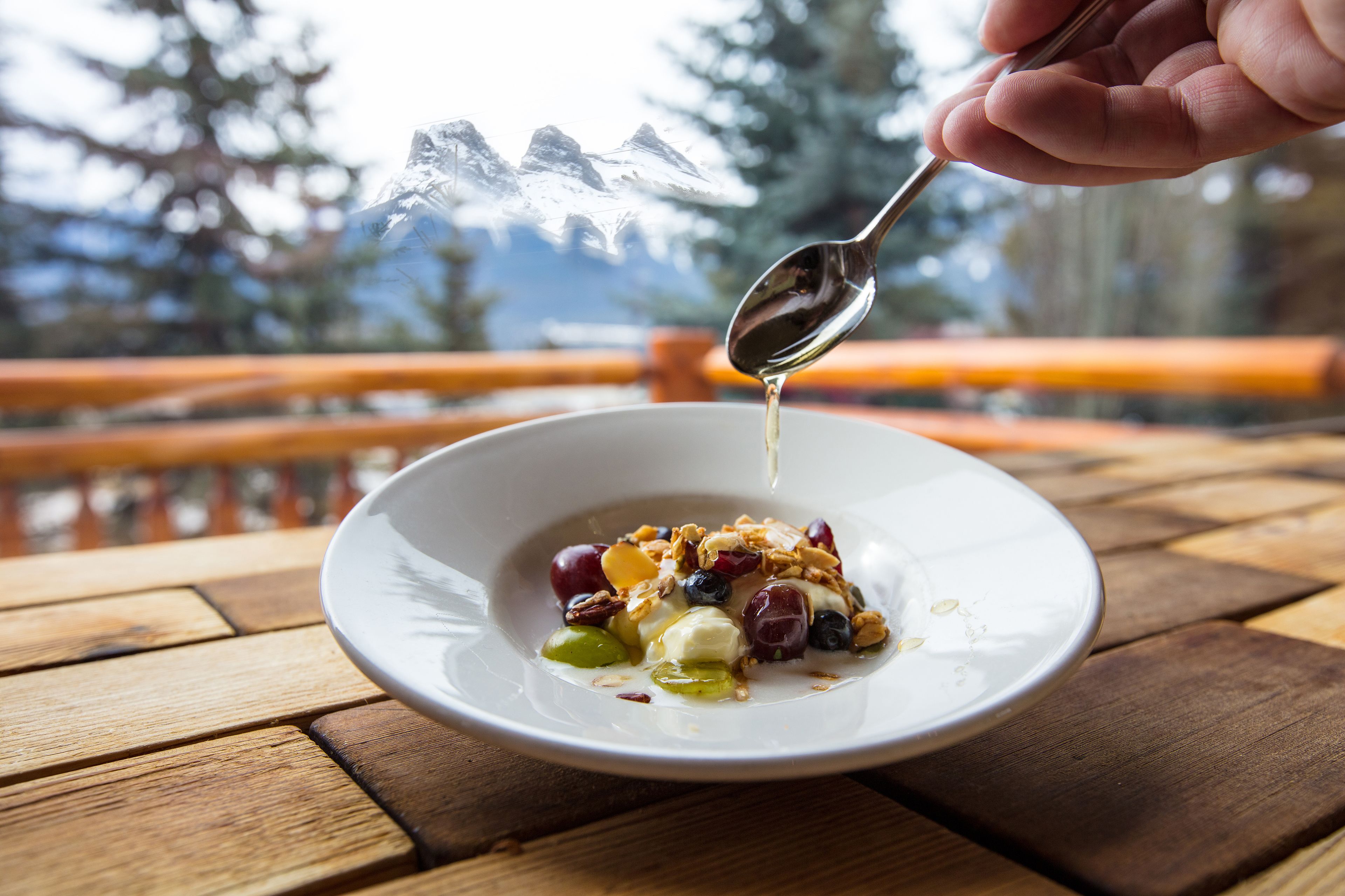 A bowl of yogurt with fruits and granola on a wooden table with a mountain view in the background.